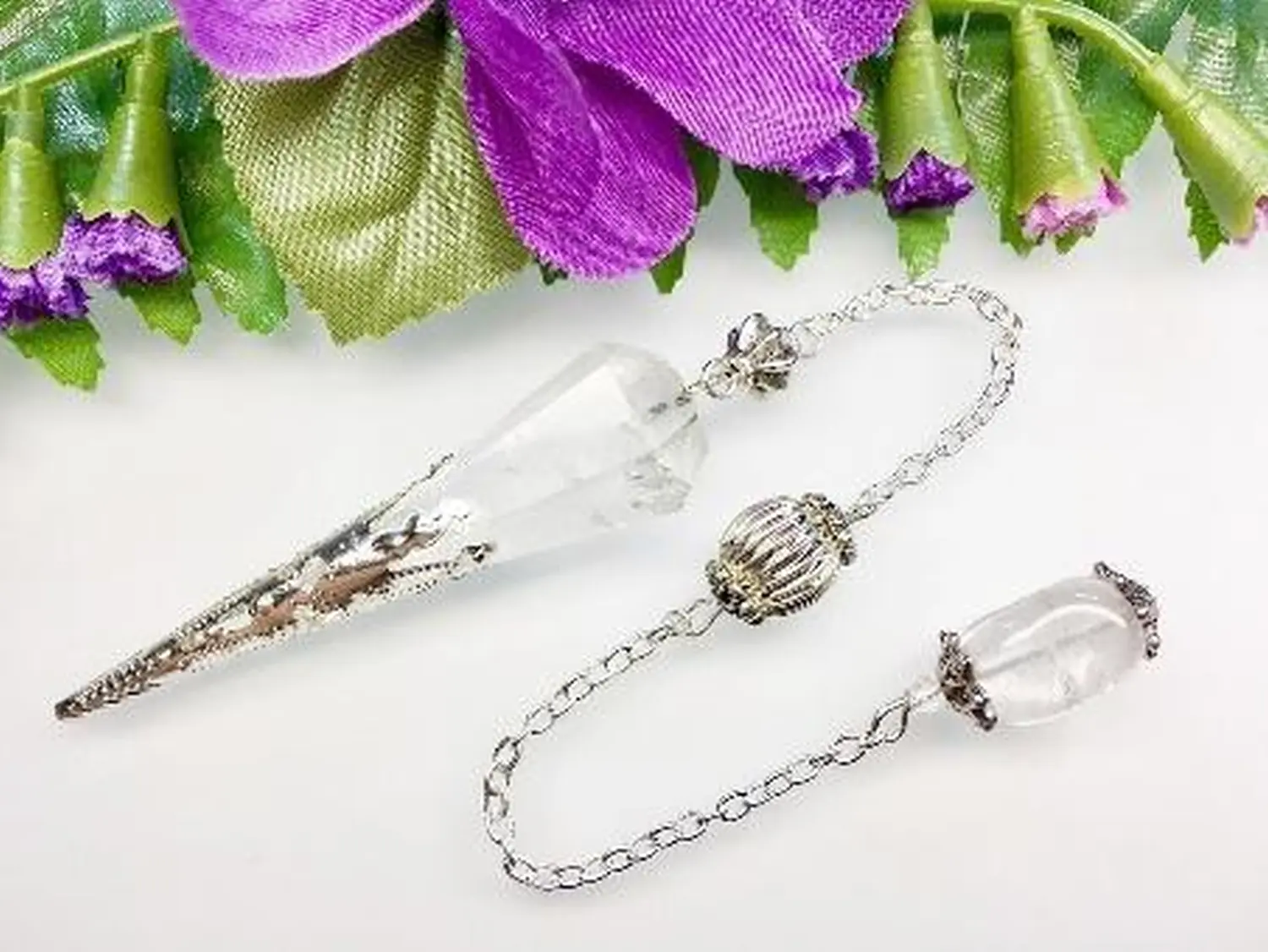enHealing - Energetischen Praxis, persönlichen Entwicklung und Vitalstoffberatung - Ein Kristallpendel mit silberner Kette und Perle liegt auf einer weißen Fläche neben lila Blüten.